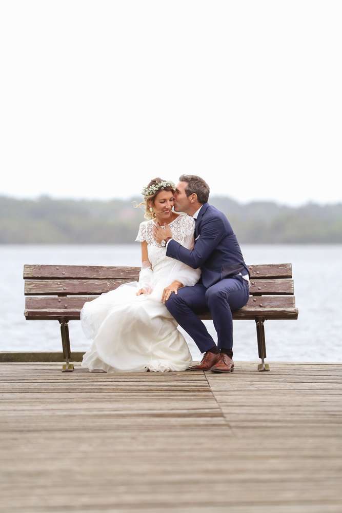 les mariés sont au bout d'un ponton d'un lac assis sur un banc, le marié embrasse sa femme dans le cou